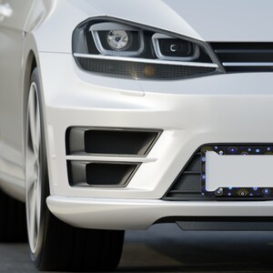 May include: A white car with a black license plate frame featuring a blue and yellow evil eye pattern. The frame is attached to the front bumper of the car.