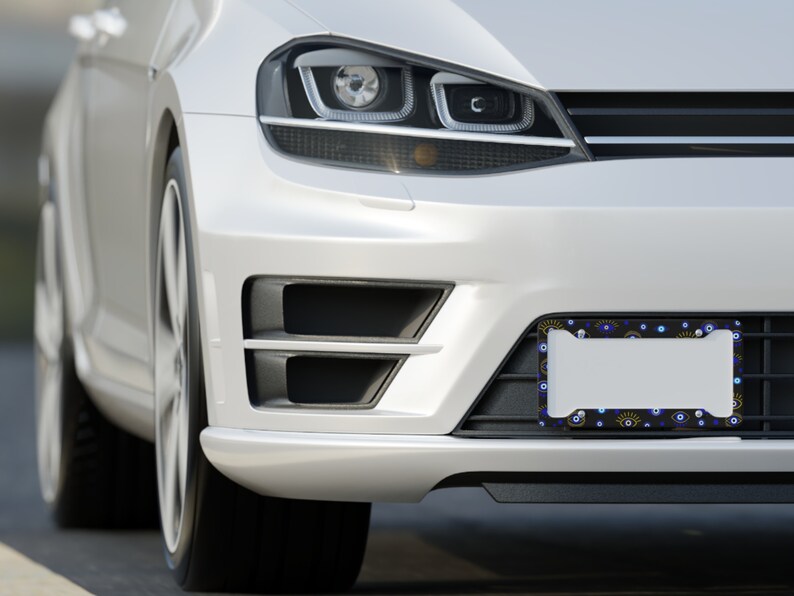 May include: A white car with a black license plate frame featuring a blue and yellow evil eye pattern. The frame is attached to the front bumper of the car.