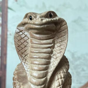 Könnte beinhalten: Eine detaillierte Holzskulptur einer Kobra, geschnitzt mit komplizierten Schuppen und Gesichtszügen. Die Kobra wird in einer erhobenen, aufmerksamen Pose dargestellt, die die Handwerkskunst der Holzschnitzerei zeigt. Die Skulptur ist auf einem kleinen, dunklen Holzsockel montiert.