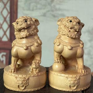 May include: Two wooden Foo Dog statues. Each statue is carved from light-colored wood and features a lion-like creature with detailed facial features, a decorative collar, and a base with ornate carvings. One holds a ball, and the other has a small cub.