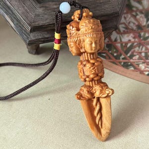 May include: A carved wooden pendant with multiple faces and a blade design. The pendant is attached to a dark brown cord with a white bead and yellow and red accents. The wood has a natural, light brown colour.