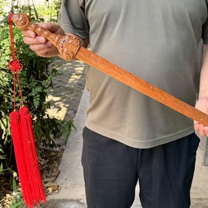 Può includere: Una spada di legno con intagli intricati sull'impugnatura e una lama lunga e piatta. La spada è di colore marrone chiaro e ha un nappa rossa attaccata. La spada è tenuta da una persona.