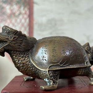 以下が含まれることがあります： 龍の頭と亀の体を持つ神話上の生き物のブロンズ色の金属製フィギュア。甲羅には中国語の文字が刻まれており、首と脚には質感のある鱗があり、口は開いています。