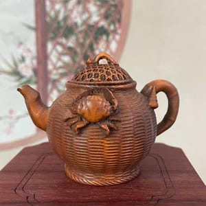 May include: A detailed wooden teapot with a crab design. The teapot is brown with a textured, woven appearance. The lid has a decorative pattern, and the handle is curved. The teapot is sitting on a dark wooden surface.