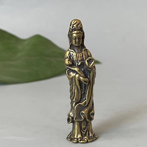 Può includere: Piccola statua in ottone in stile antico di una figura in piedi con abiti ornati e un'espressione serena. La figura tiene un piccolo oggetto in una mano. La statua è su una superficie bianca.