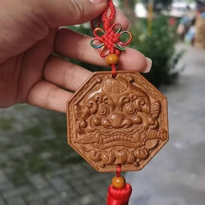 Puede incluir: Un colgante de madera marrón, octogonal, con un diseño de rostro tallado, suspendido por un cordón rojo con un nudo decorativo y una borla. El colgante está sostenido por una mano.