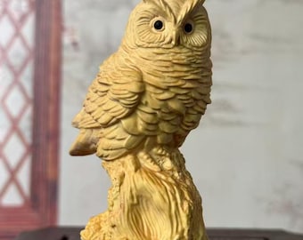 May include: A light tan wooden owl figurine perched on a textured tree stump. The owl features detailed carved feathers, pointed ear tufts, and two small, dark, circular eyes. The entire sculpture is crafted from a single piece of light-colored wood.