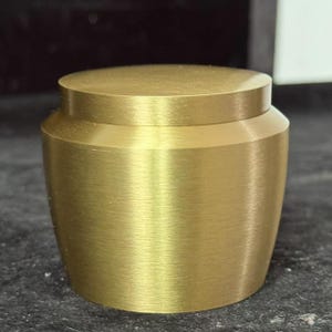 May include: A small, cylindrical, gold-colored container with a lid. The container has a brushed metal finish and a slightly tapered body. The lid sits flush on top, creating a clean, minimalist design. The container is likely used for storage or decorative purposes.