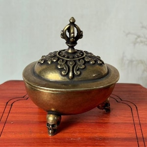 Peut inclure: Un brûleur d'encens en bronze avec un couvercle décoratif et trois pieds en forme de crâne. Le couvercle est orné de motifs floraux complexes et d'un fleuron stylisé. Le brûleur est posé sur une surface en bois rouge.