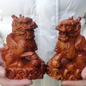 May include: Two intricately carved wooden statues of mythical creatures, possibly dragons or lions, in a reddish-brown hue. Each statue features detailed wings, armor, and a fierce expression. The figures are posed on decorative cloud-like bases.