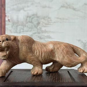 Puede incluir: Escultura de madera marrón claro que representa un tigre en posición de acecho. La boca del tigre está abierta, revelando los dientes, y su pelaje está texturizado. La escultura se exhibe sobre una base de madera oscura.