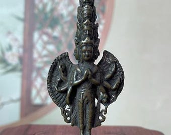 May include: A bronze-colored statue of a deity with multiple arms and a layered headpiece. The figure stands on a detailed base, with wings and a serene expression. The statue is displayed on a wooden surface.