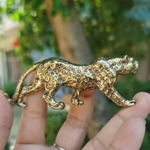 May include: A golden-colored leopard figurine in a walking pose. The spotted cat sculpture is made of metal and has a detailed texture. The figurine is held in a hand against a blurred green background.