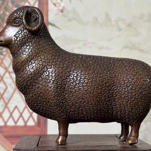 Puede incluir: Una figura de carnero color bronce que presenta un pelaje texturizado detallado con patrones de remolinos repetitivos en todo el cuerpo. La escultura muestra al animal en posición de perfil, con cuernos curvados y cuatro patas robustas.