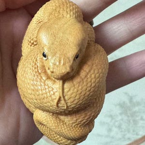 Könnte beinhalten: Ein detaillierter Holzanhänger in Form einer zusammengerollten Schlange. Der Anhänger hat eine hellbraune Farbe mit sichtbaren Schuppen und einem detaillierten Kopf. Die Schlange ist an einer schwarzen Perlenkette befestigt.