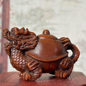 Peut inclure: Sculpture en bois détaillée d'une créature mythique, un hybride dragon-tortue. La sculpture marron présente des écailles, des griffes et une carapace en forme de théière. La sculpture est exposée sur une surface rouge.