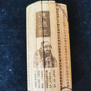 Könnte beinhalten: Ein antikes Bambusbuch mit chinesischen Schriftzeichen und einer Illustration eines Mannes. Das Buch ist mit einem braunen Faden gebunden und hat eine gebogene Form. Der Einband zeigt den Titel "道 德 經".