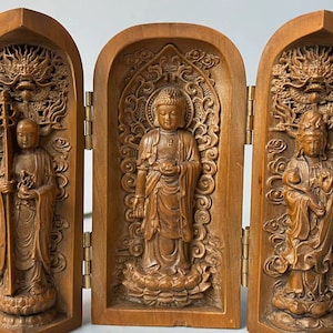 Puede incluir: Un tríptico de madera con figuras budistas talladas. El panel central muestra un Buda de pie, flanqueado por figuras que sostienen objetos simbólicos. La madera tiene un tono cálido y natural, y las tallas son detalladas.