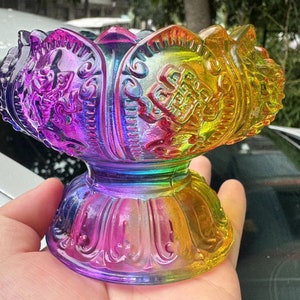 May include: A multi-coloured, iridescent lotus flower-shaped decorative object. The object has a pedestal base and is held in a hand. The colours transition from purple to yellow, with intricate embossed details.