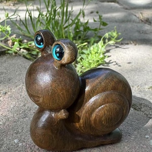 May include: A brown wooden snail ornament with a polished finish. The snail features large eyes with blue irises. The sculpture is set on a stone surface, with green plants in the background. The snail's body has carved details.