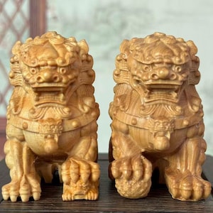 May include: Two intricately carved wooden guardian lion statues. The statues are light brown, with detailed features including manes, facial expressions, and paws. They are positioned side-by-side, showcasing the craftsmanship of the wood carving.