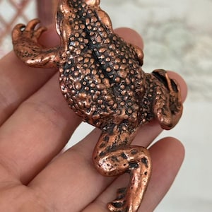May include: A detailed copper-colored frog figurine with a textured surface. The frog is posed with its legs extended and arms raised, showcasing its intricate design. The figurine is held in a hand, highlighting its size and detail.