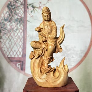 May include: A wooden sculpture of a seated figure, possibly a deity, with flowing robes and serene expression. The figure holds an object and is seated on a stylized lotus flower base. The sculpture is carved from light-colored wood, showcasing the natural grain.