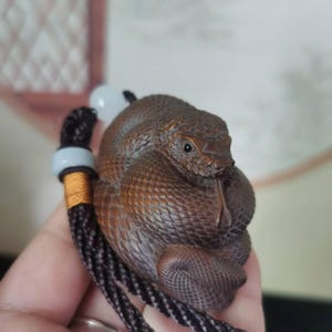 Puede incluir: Una detallada talla de madera marrón de una serpiente enroscada con la lengua extendida. La serpiente está unida a un cordón trenzado negro y dorado con una cuenta blanca y gris.