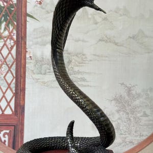 May include: A bronze-colored cobra statue with an open mouth, poised to strike. The snake's body is intricately detailed, with scales visible. The statue is mounted on a small, dark wooden base, showcasing a detailed sculpture.