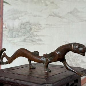 Könnte beinhalten: Eine bronzefarbene Skulptur einer stilisierten Katze, möglicherweise eines Leoparden oder Jaguars, in einer dynamischen Pose. Die Skulptur steht auf einem dunklen Holzsockel, wobei der Körper des Tieres gewölbt und die Beine ausgestreckt sind. Der Hintergrund zeigt eine gemalte Landschaft.