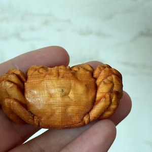 Può includere: Piccola statuetta di granchio in legno intagliato con precisione. Il granchio è di un caldo color marrone chiaro, con chele e segmenti del corpo dettagliati. L'intaglio mette in risalto la venatura naturale del legno.