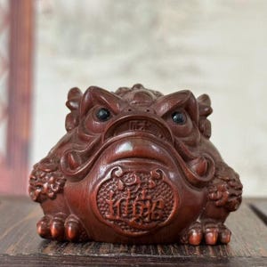 May include: A detailed, reddish-brown wooden frog sculpture. The frog has a detailed carved design, with prominent eyes and a textured body. The frog's belly features an ornate, circular carving with Chinese characters. The sculpture is displayed on a wooden surface.