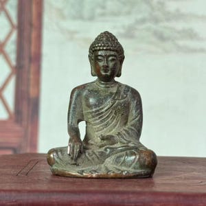 Può includere: Piccola statua di Buddha color bronzo, in posizione di meditazione seduta. La figura ha un'espressione serena, lineamenti dettagliati ed è drappeggiata in una veste. La statua è su una superficie di legno scuro.