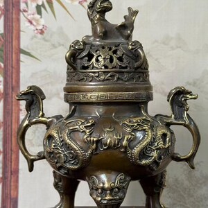 May include: A bronze-coloured metal incense burner featuring a lid topped with a mythical creature figure. The body is decorated with embossed dragon motifs and two side handles shaped like dragon heads, supported by three ornate legs.