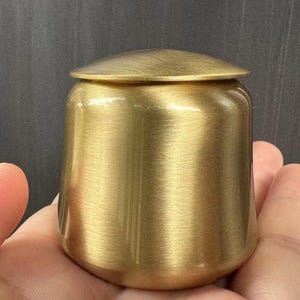May include: A small, cylindrical brass container with a matching lid. The container has a brushed finish, giving it a matte gold appearance. The lid sits flush on top. The container is held in a hand.