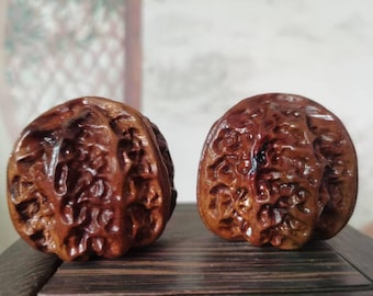 Puede incluir: Dos esculturas de madera tallada de nueces. Las esculturas son de un color marrón oscuro intenso con un acabado brillante, que muestra texturas y patrones detallados. Se encuentran una al lado de la otra sobre una superficie de madera, con un fondo borroso.