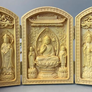 May include: A wooden triptych featuring carved figures of Buddha and other deities. The central panel depicts Buddha seated on a lotus flower, flanked by two figures. The side panels show additional figures, all in a light wood color.