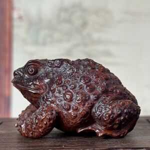 以下が含まれることがあります： 濃い茶色の木彫りのヒキガエルの置物で、表面は凹凸のある質感に仕上げられています。背中と側面には詳細な円形の模様が施されており、目と大きくはっきりとした口が特徴的な、座った姿勢の彫刻作品です。