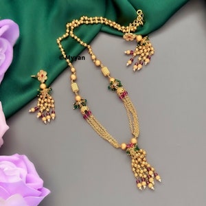 May include: A gold necklace and earrings set with a beaded tassel design. The necklace features a chain with small gold balls and green and red beads. The earrings have a flower design with a red center and a beaded tassel.