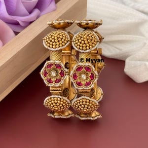 May include: A set of four gold bangles with intricate designs. Each bangle features a red flower with white pearl accents and a gold dome top. The bangles are stacked on top of each other.