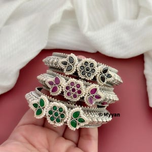 Könnte beinhalten: Vier silberne Armreifen mit floralen Designs und bunten Edelsteinen. Die Armreifen sind übereinander gestapelt und haben schwarze, rosa und grüne Edelsteine.