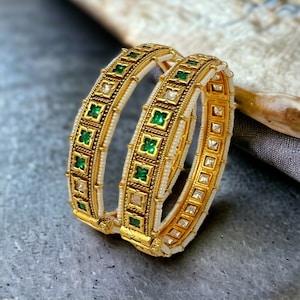 May include: Two gold bangles with intricate designs. Each bangle features a white beaded inner band, accented with square emerald green and clear crystal-like stones. The outer band is gold with detailed patterns. The bangles are displayed on a gray surface.