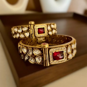 May include: Two gold bangles with intricate designs featuring small white stones and a large red gemstone in a square setting.