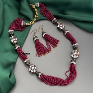 May include: A maroon beaded necklace with decorative oval-shaped beads and matching tassel earrings. The beads are accented with rhinestones and have a white and gray pattern.