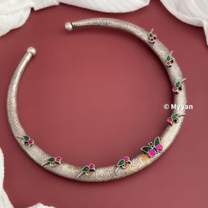 May include: A silver choker necklace with an intricate design and green and pink butterfly accents. The necklace is made of metal and has a circular shape.