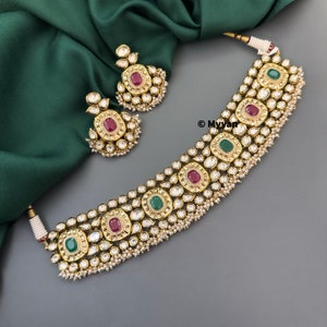 May include: A gold choker necklace with green and red stones, and matching earrings. The necklace is decorated with small white pearls.