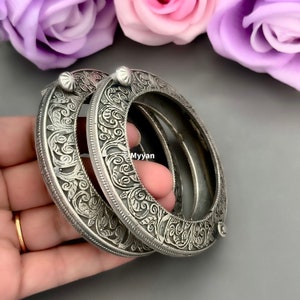 May include: Two silver-toned metal bangle bracelets with intricate floral designs. The bracelets are oval-shaped and have a slightly raised edge.