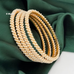 May include: A set of gold bangles adorned with small, creamy-white pearls. The pearls are intricately woven around the gold bands, creating a textured, rope-like appearance. The bangles are displayed on a dark green, silky fabric.