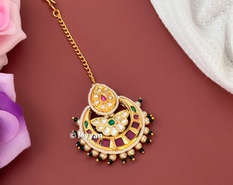 May include: Gold-toned Indian headpiece with a crescent-shaped design. The headpiece features a central teardrop shape and is adorned with red, green, and white gemstones, surrounded by small pearls. A delicate gold chain extends to a hook.