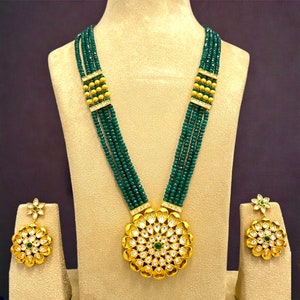 May include: A gold necklace with a large, round pendant featuring a floral design with white and green stones. The necklace is made of multiple strands of green beads and gold accents. Matching earrings with a similar floral design are also included.